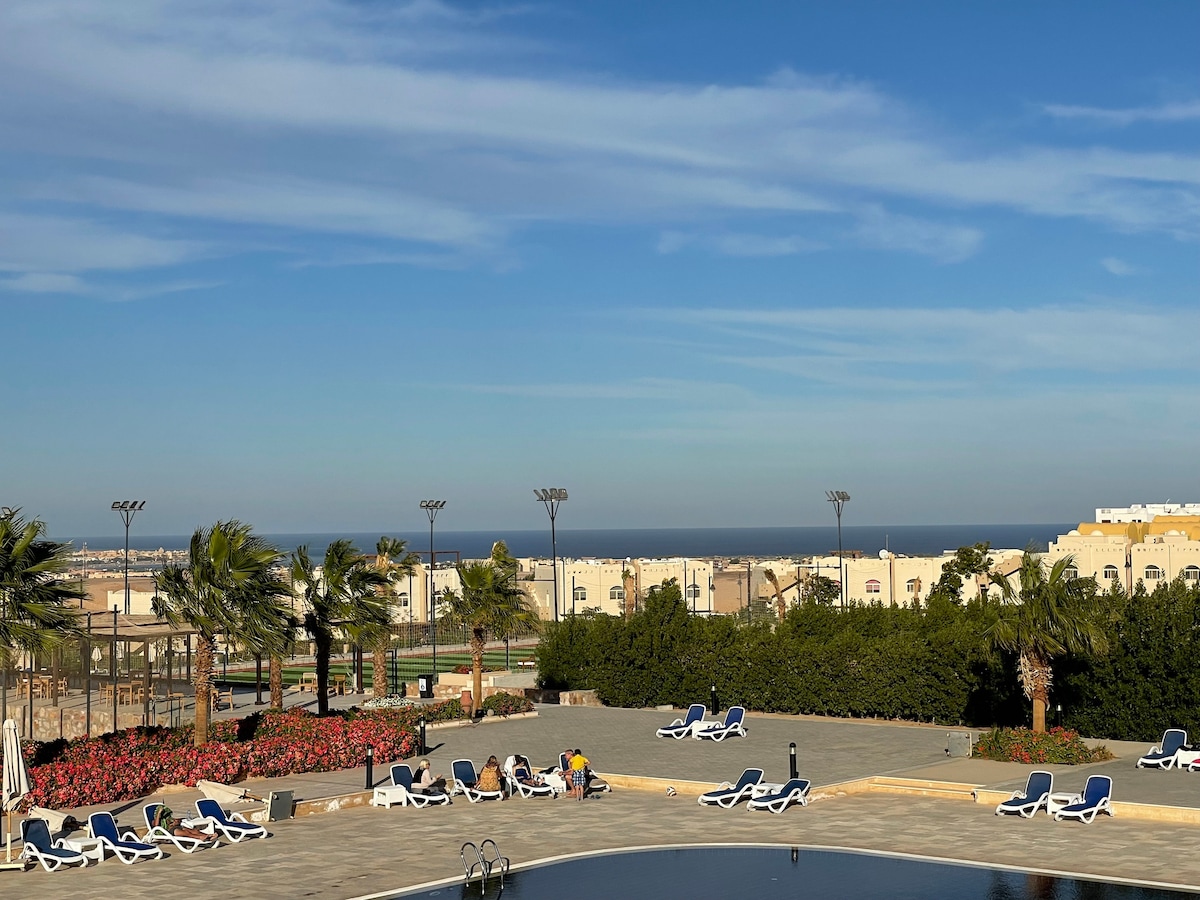 A serene outdoor area features numerous lounge chairs arranged around a swimming pool, set against a backdrop of a vibrant floral garden and palm trees. A clear blue sky stretches overhead, while the distant coastline is visible beyond the landscaped grounds.