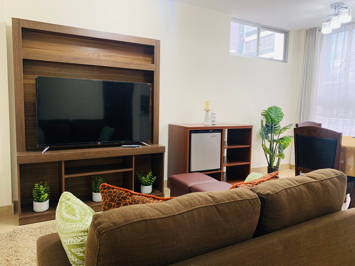 A welcoming living area is highlighted by a large television mounted on a wooden shelf. A sectional sofa with colorful cushions faces the TV, while a small refrigerator is placed in a nearby cabinet. Natural light enters through large windows, complemented by indoor plants.