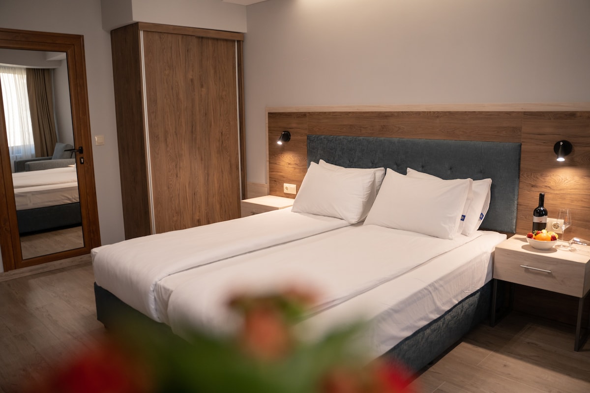 A well-appointed guest room features two spacious beds with crisp white linens. A wooden accent wall adds warmth, and a mirror reflects the light from the window. A small table displays a bottle of wine and glasses, contributing to a relaxed atmosphere.