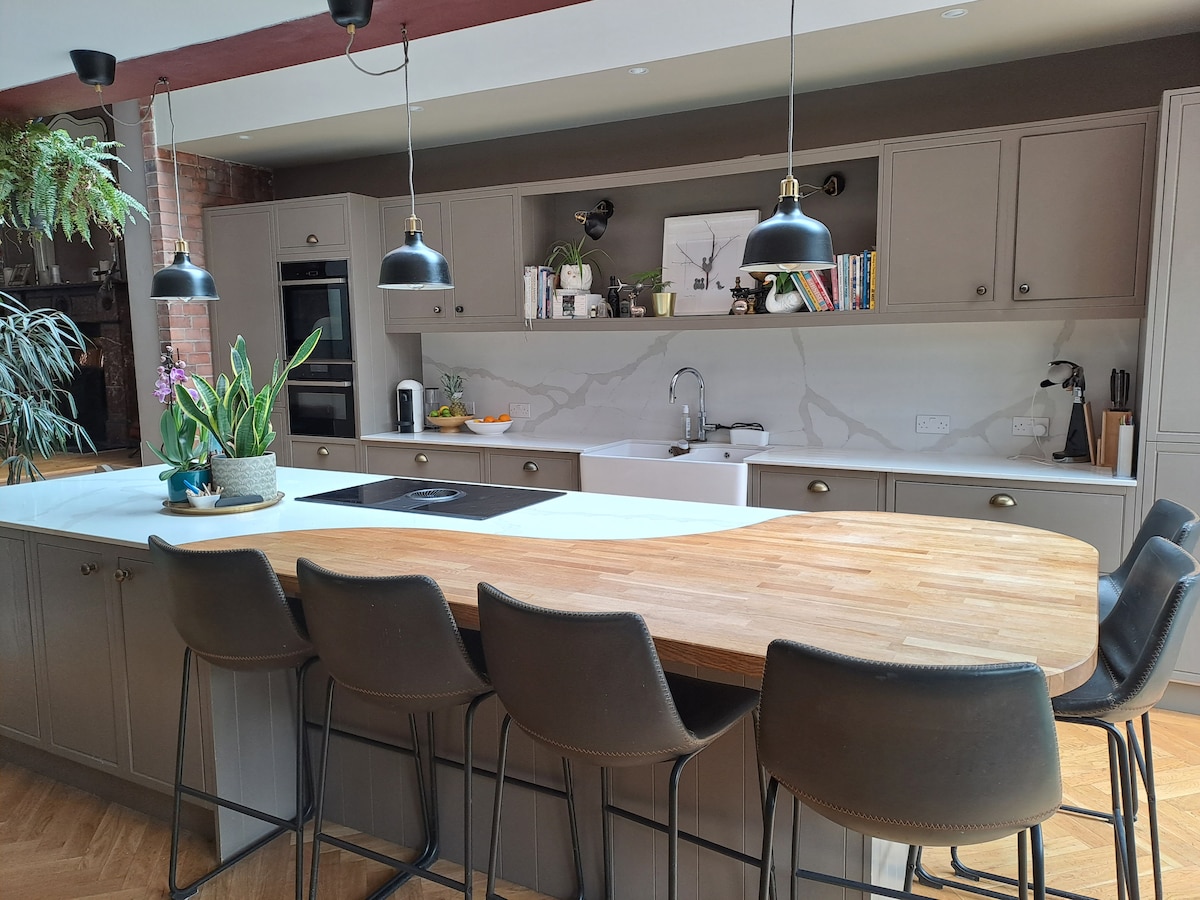 A spacious kitchen features a large island with a curved wooden countertop, surrounded by several bar stools. Modern cabinetry in muted tones provides ample storage, while overhead pendant lights offer focused lighting. A plant adds a touch of greenery, contributing to a bright and inviting atmosphere.