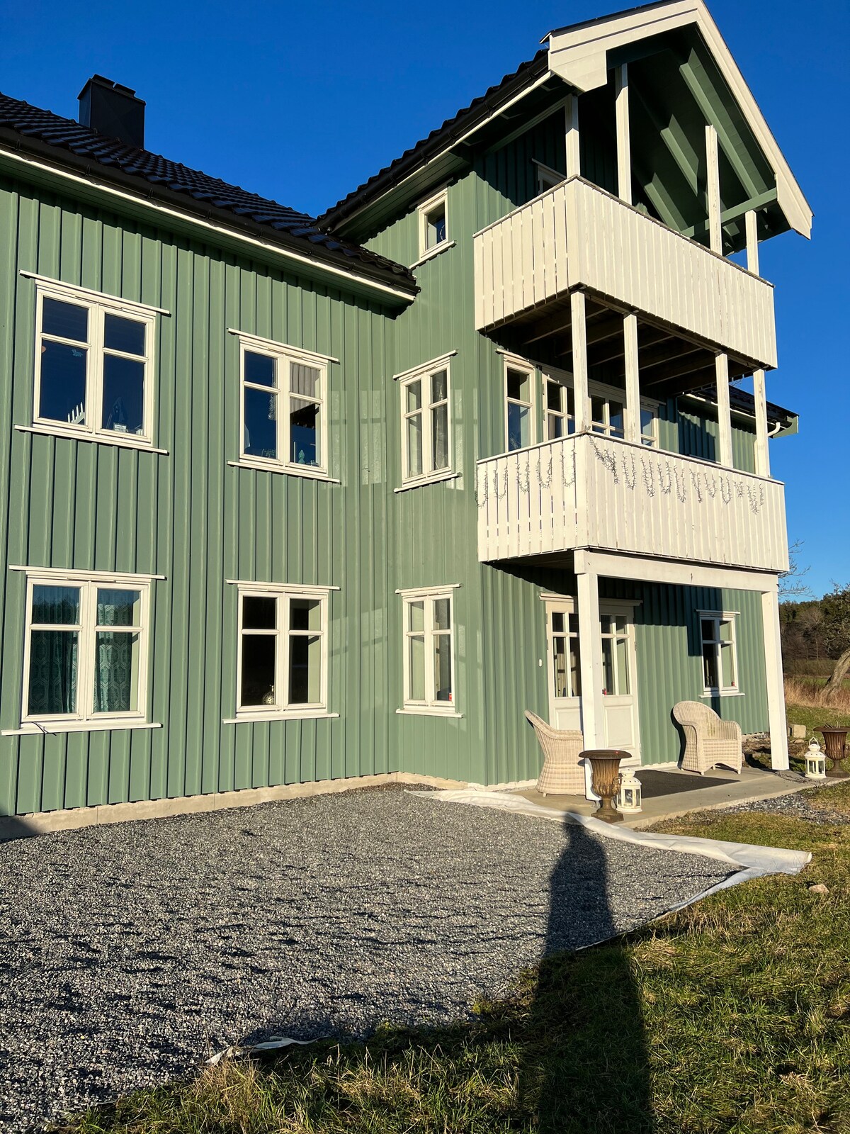 A green, two-story house is shown, featuring a prominent balcony above the entrance. Large windows allow for natural light, while outdoor seating is positioned nearby on the gravel surface. The surrounding area includes grass and a clear blue sky.
