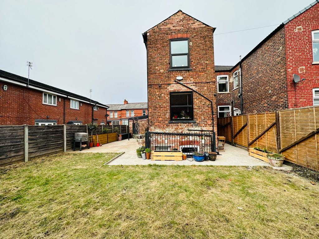 A spacious garden area features a paved patio with seating arrangements and a barbecue grill. The brick facade of the apartment stands prominently against a neatly enclosed outdoor space, bordered by wooden fencing and landscaped with potted plants. A well-maintained lawn occupies the center area.