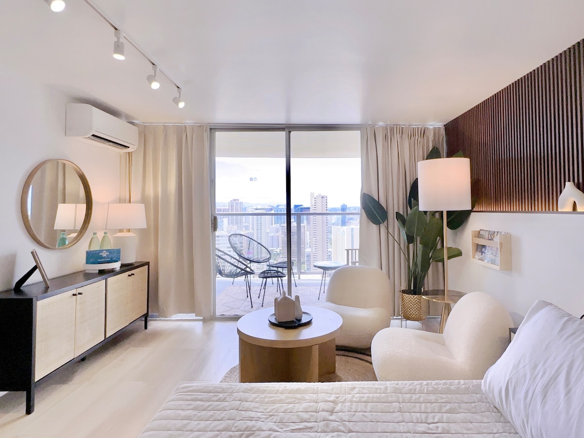 A spacious living area features a king size bed with a soft white blanket, complemented by a round coffee table and two boucle fabric chairs. Large windows allow natural light to fill the room, revealing views of the Waikiki skyline and hinting at partial ocean vistas.