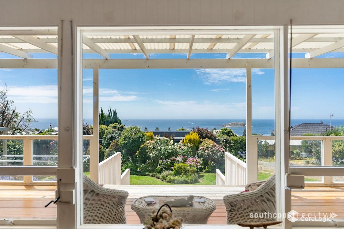 A wide, open view showcases vibrant gardens and the ocean beyond, framed by a large glass window. Comfortable seating arrangements invite relaxation, while the covered deck provides shade. The surrounding greenery enhances the serene environment, offering a peaceful retreat.