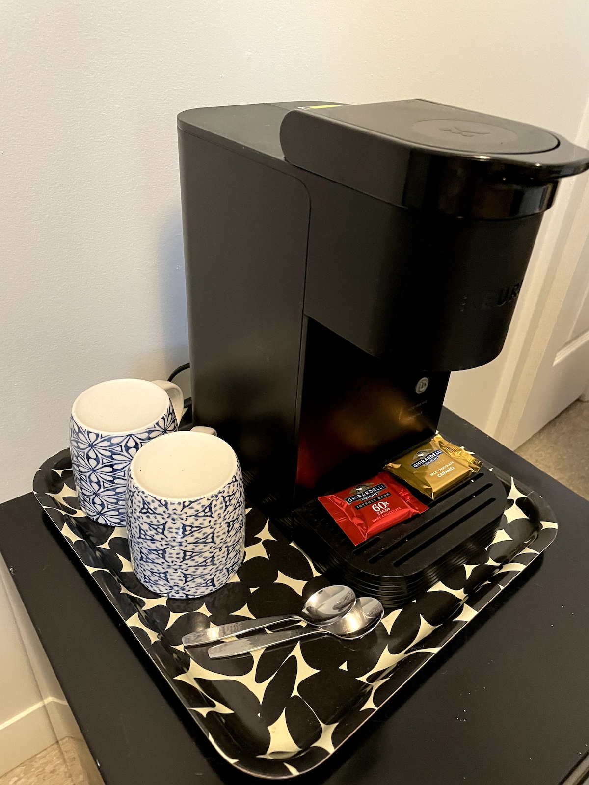 A coffee station is displayed on a tray featuring a black coffee maker beside two patterned cups. Two packets of coffee and sugar are neatly arranged, alongside two teaspoons, presenting an inviting setup for morning beverages.