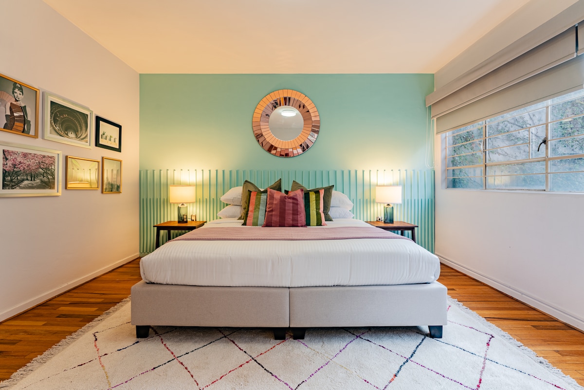A relaxing bedroom features a spacious bed with crisp linens and a variety of colorful accent pillows. The room is adorned with modern art on one wall and has a round mirror above the headboard. Natural light enters through a large window, complemented by soft, neutral tones.