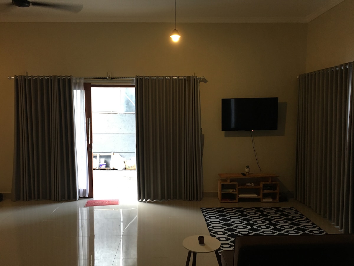A spacious living room area features a large television mounted on the wall. Light-colored walls are complemented by thick curtains that frame a glass door leading to an outdoor space. A patterned rug lies on the tiled floor, while bedside tables are positioned near seating arrangements.