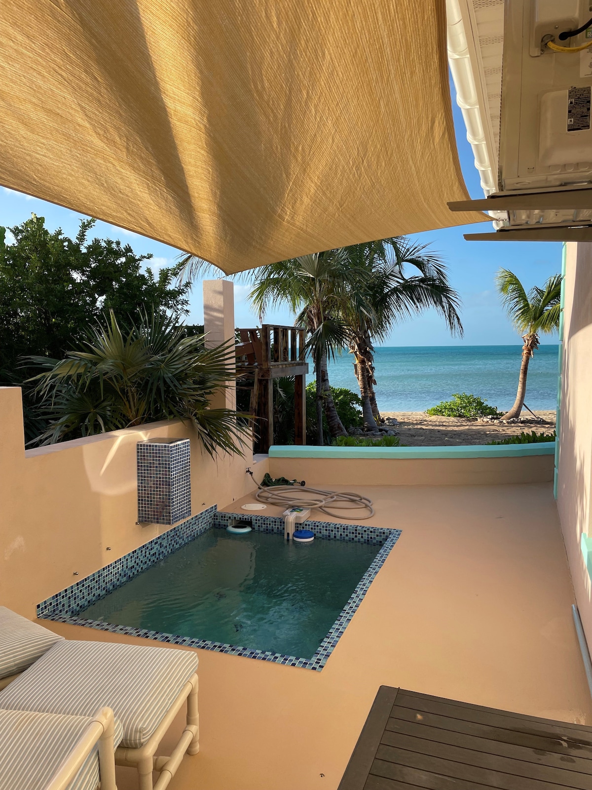 A private outdoor area features a small pool surrounded by a tiled border. Tall palm trees and a glimpse of the beach create a serene backdrop. Comfortable seating is situated nearby, with light fabric overhead providing shade from the sun.