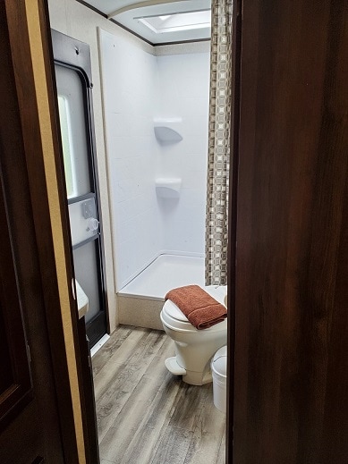 A compact bathroom is showcased, featuring a shower stall with built-in shelves and a light-colored shower curtain. A towel is neatly placed on the toilet tank, while the floor displays a wood-like finish, contributing to the modern aesthetic.