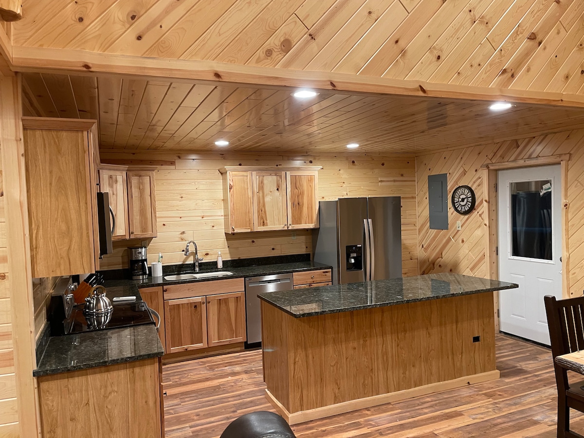 A spacious kitchen is highlighted by warm wooden cabinetry and a large dark granite countertop. Stainless steel appliances are integrated seamlessly, and soft lighting illuminates the space. Open shelving adds an inviting touch, while wooden accents complement the natural warmth of the pine walls.
