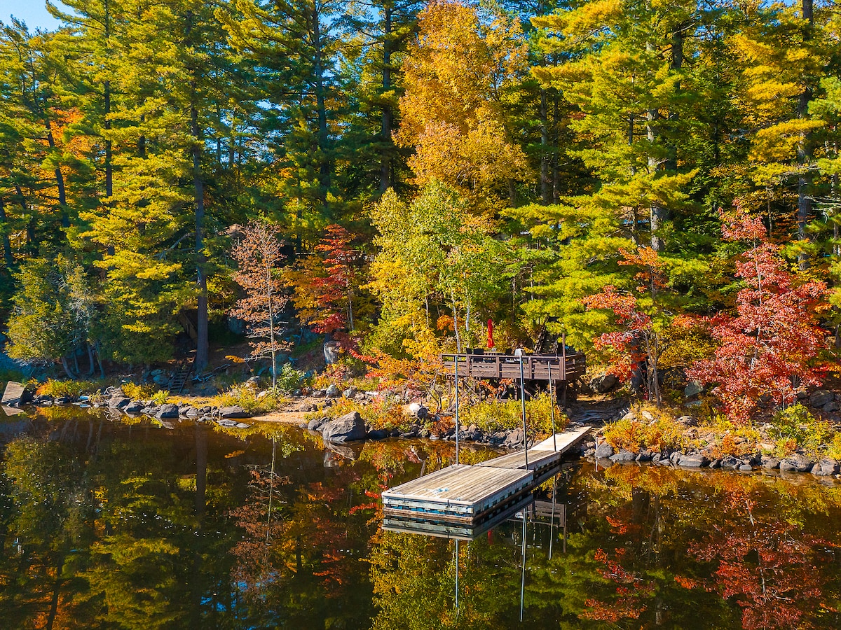A serene dock extends over calm waters, surrounded by vibrant autumn foliage. Trees display hints of red, orange, and yellow, reflecting on the lake's surface. The tranquil scene invites relaxation and enjoyment of nature.