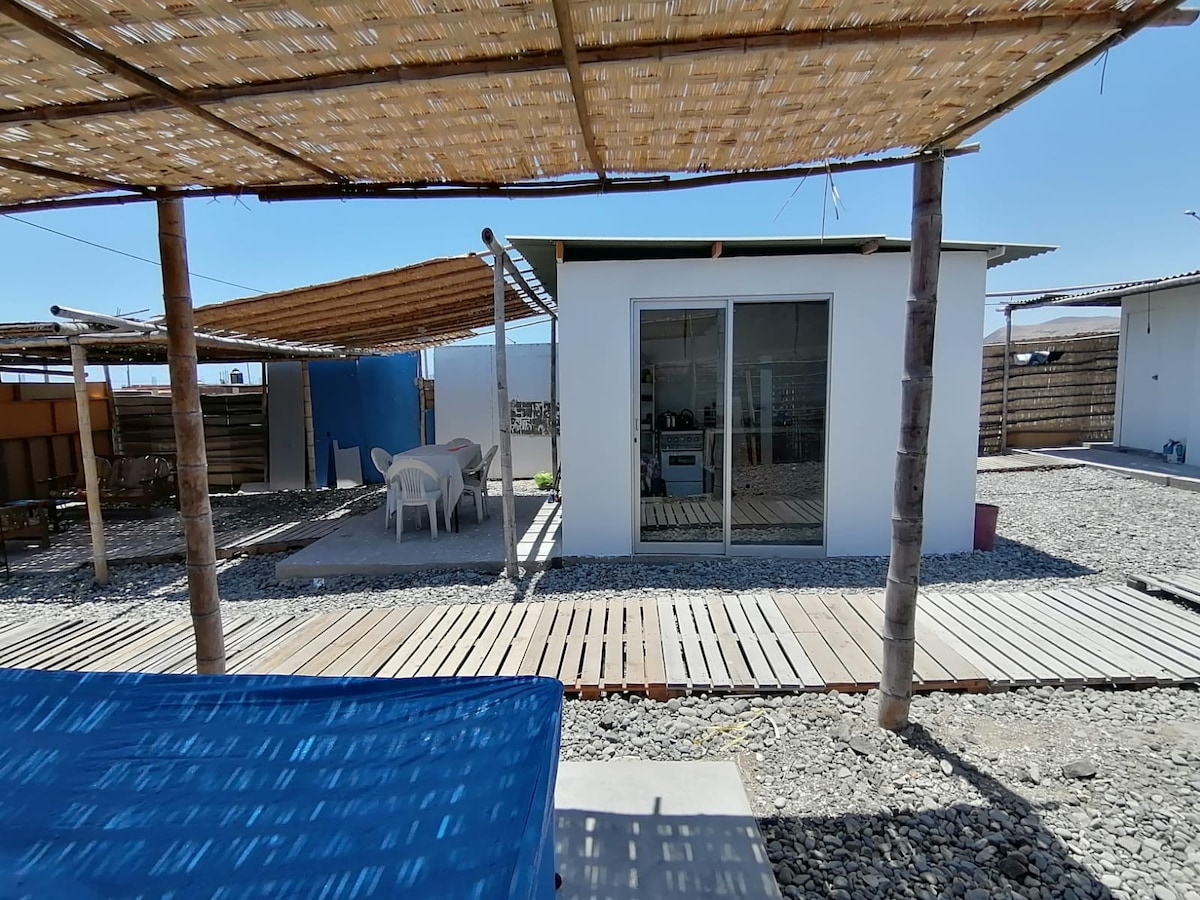 A modern structure is shown with large sliding glass doors, providing a view of the outdoor area. A wooden deck leads to a seating area with white chairs. Palm fronds create shade above, and a blue tarp partially covers the view at the foreground.