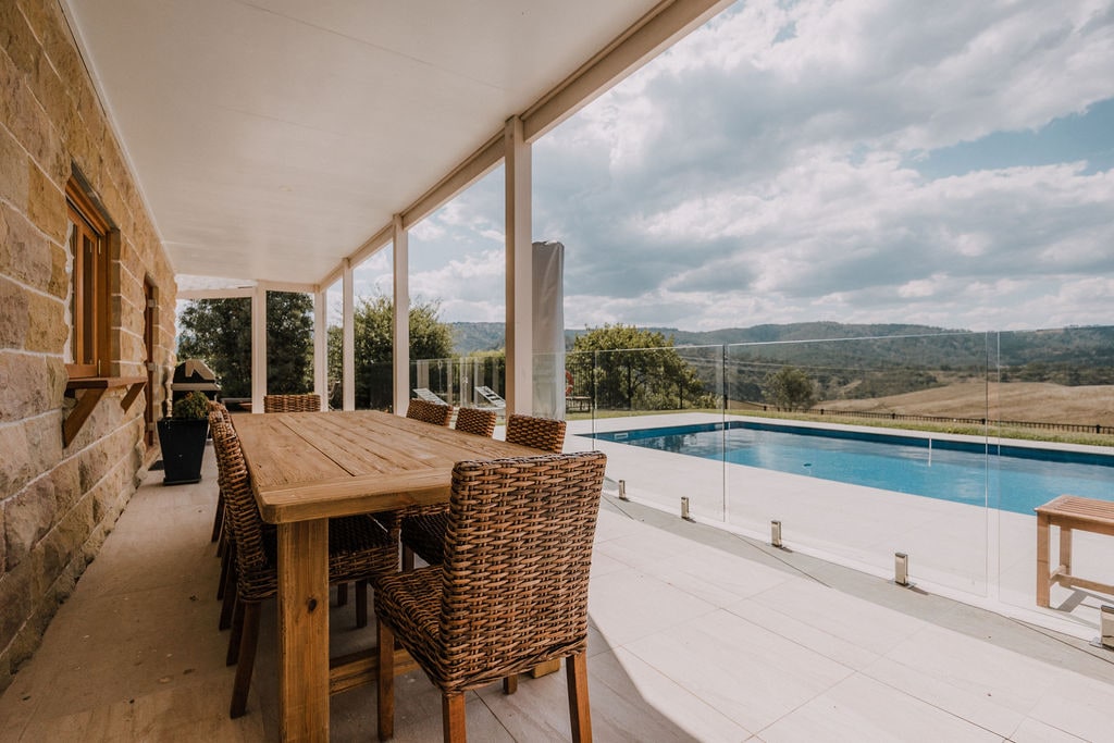 An expansive outdoor dining area features a large wooden table surrounded by woven chairs. The space is shaded and offers views of the pool and the surrounding hills, with glass panels enhancing the connection to nature.