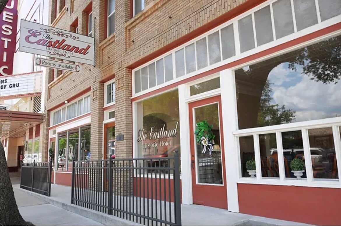 The exterior of the Eastland Historic Hotel displays a warm brick facade and large windows. The entrance is framed by decorative plants and signage, creating an inviting first impression. A black wrought-iron fence lines the front, enhancing the historic charm of the surroundings.