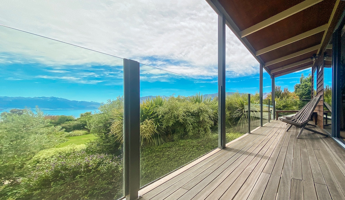 A spacious deck features a comfortable lounge chair, overlooking lush greenery and panoramic views of Lake Hawea and the surrounding mountains. Glass panels provide unobstructed views, while the wooden floor complements the natural setting.