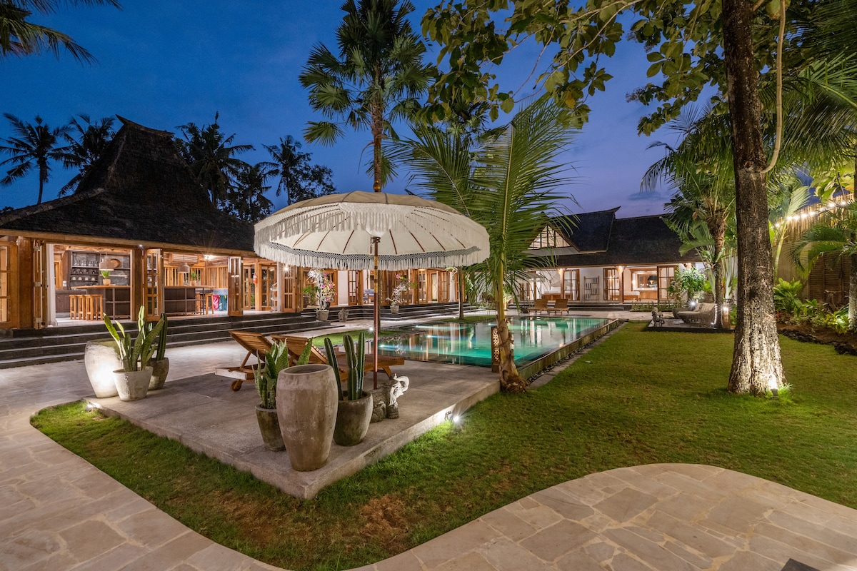 A tranquil evening scene captures the spacious outdoor area of the villa, featuring a large pool surrounded by lush tropical gardens. Comfortable loungers are placed under a shaded sun umbrella, with decorative pots enhancing the serene environment.