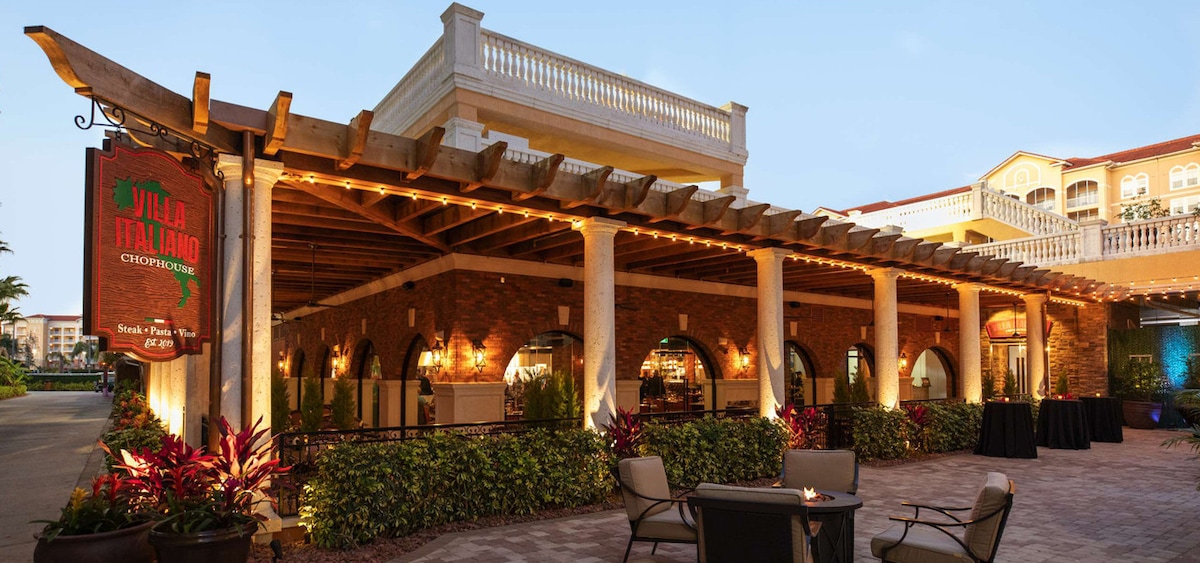 An outdoor dining area is highlighted by decorative lighting and lush greenery. The building features a classic architectural style with arched doorways and a wooden pergola above the patio. Several seating areas with chairs are prominent, inviting guests to relax in a charming setting.