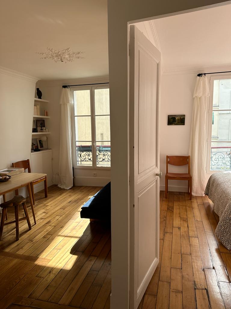 A well-lit interior space is captured, showcasing a cozy bedroom and a living area. Natural light filters through large windows, illuminating the wooden flooring. A small dining table with chairs is visible, along with a comfortable seating area, creating an inviting atmosphere.