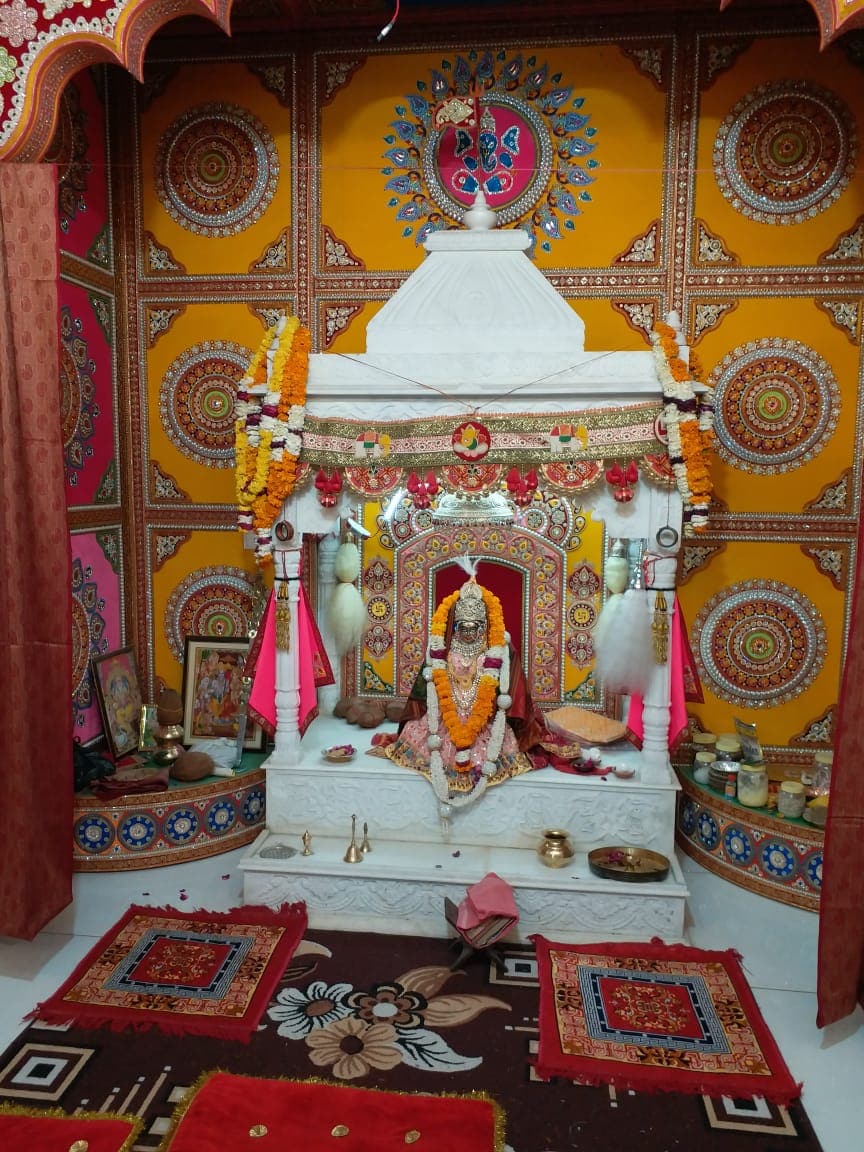 A dedicated prayer area features a white marble altar adorned with floral garlands and incense holders. Intricate wall designs in vibrant colors surround the altar. Traditional rugs in warm tones add a cozy touch, while idols and decorative items are arranged thoughtfully.