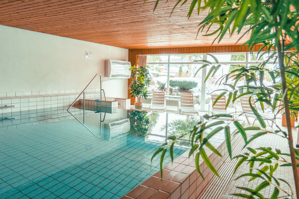 An indoor swimming pool is surrounded by a wooden ceiling and potted plants, creating a serene atmosphere. Sunlight filters through large windows, reflecting off the clear water. Comfortable loungers are positioned nearby, inviting relaxation after a refreshing swim.