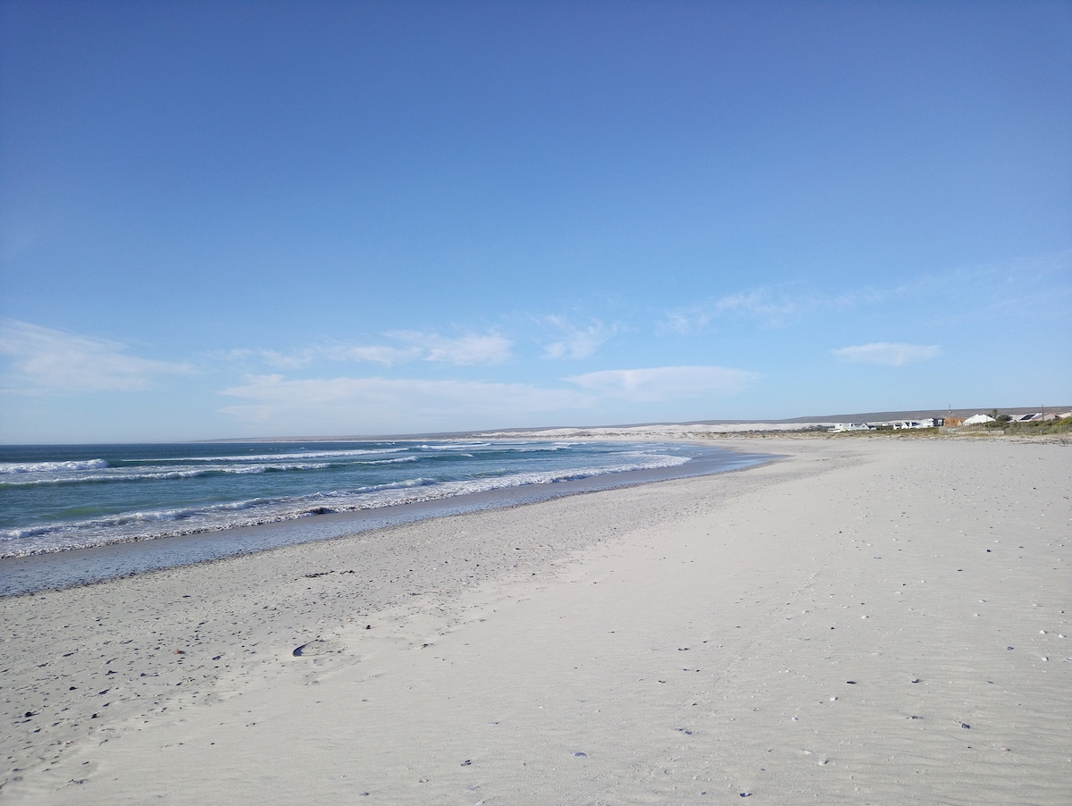 A serene beach scene features a wide stretch of soft, white sand framed by gentle waves of a calm sea. The clear blue sky is dotted with a few soft clouds, creating a tranquil atmosphere that invites relaxation and reflection.