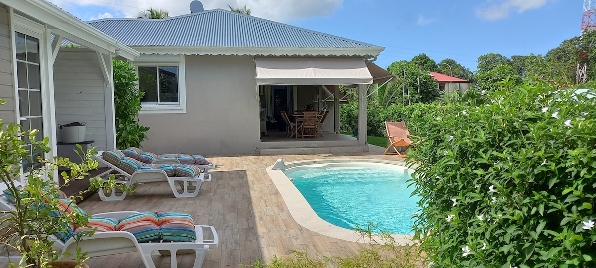 An outdoor area features a swimming pool surrounded by a tiled deck. Colorful lounge chairs are positioned beside the pool, offering a relaxing space. A covered patio with dining furniture is visible, and lush greenery adds to the serene environment.