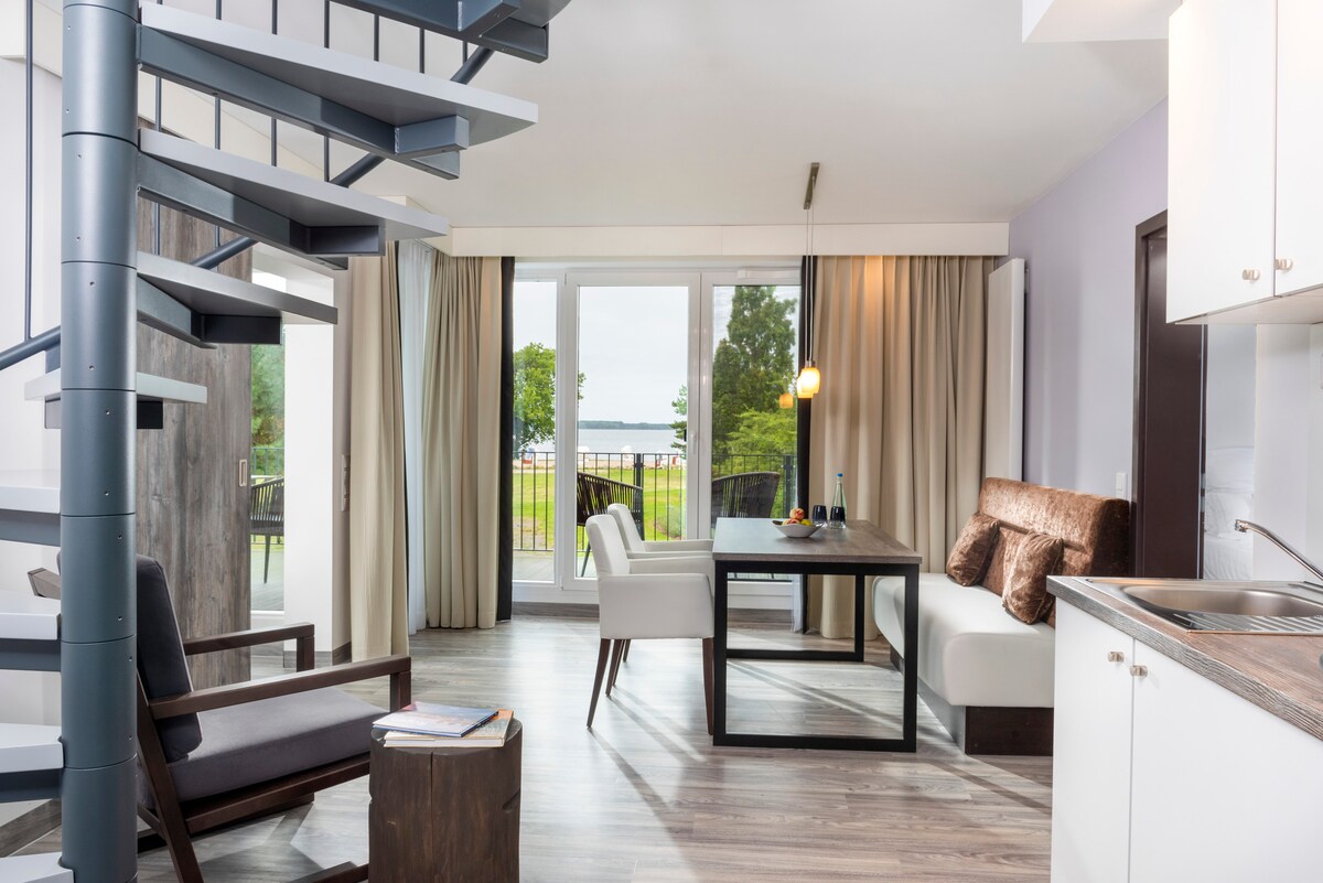 An open-plan living area features a sleek spiral staircase leading to an upper level. A dining table is set beside large glass doors, offering views of the outdoors. Comfortable seating is arranged nearby, complemented by soft natural light filtering through the curtains.