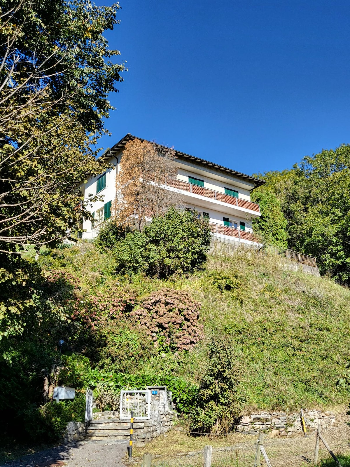 A three-story house is positioned on a hillside, surrounded by greenery and flowering plants. The building features multiple balconies and large windows, while the blue sky adds brightness to the scene, enhancing the natural setting.