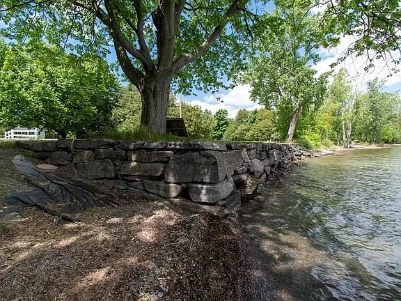 A stone retaining wall lines the water's edge, gently sloping towards the lake with a mix of sandy and rocky shore. Lush green trees provide shade nearby, and sunlight reflects off the calm water, creating a serene outdoor setting.
