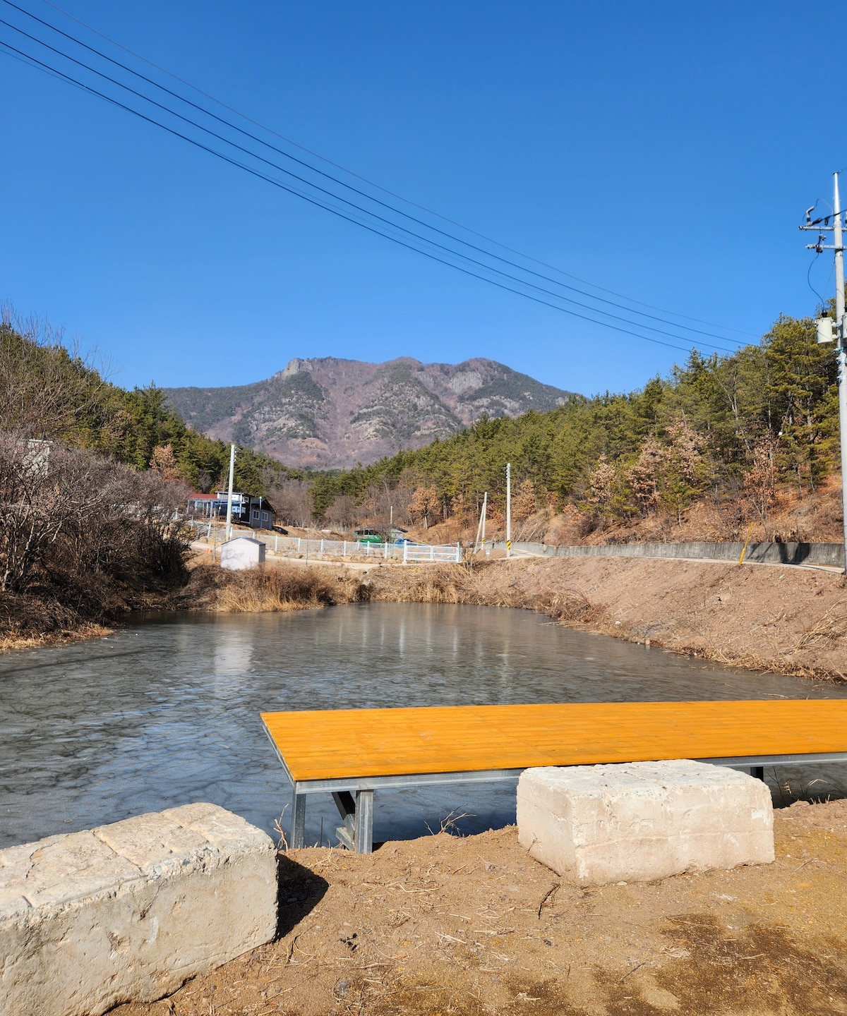 A serene body of water is shown, framed by a wooden dock extending over the surface. The surrounding landscape features trees and a mountain in the background, under a clear blue sky. Power lines are visible along the edge, adding to the scenic view.