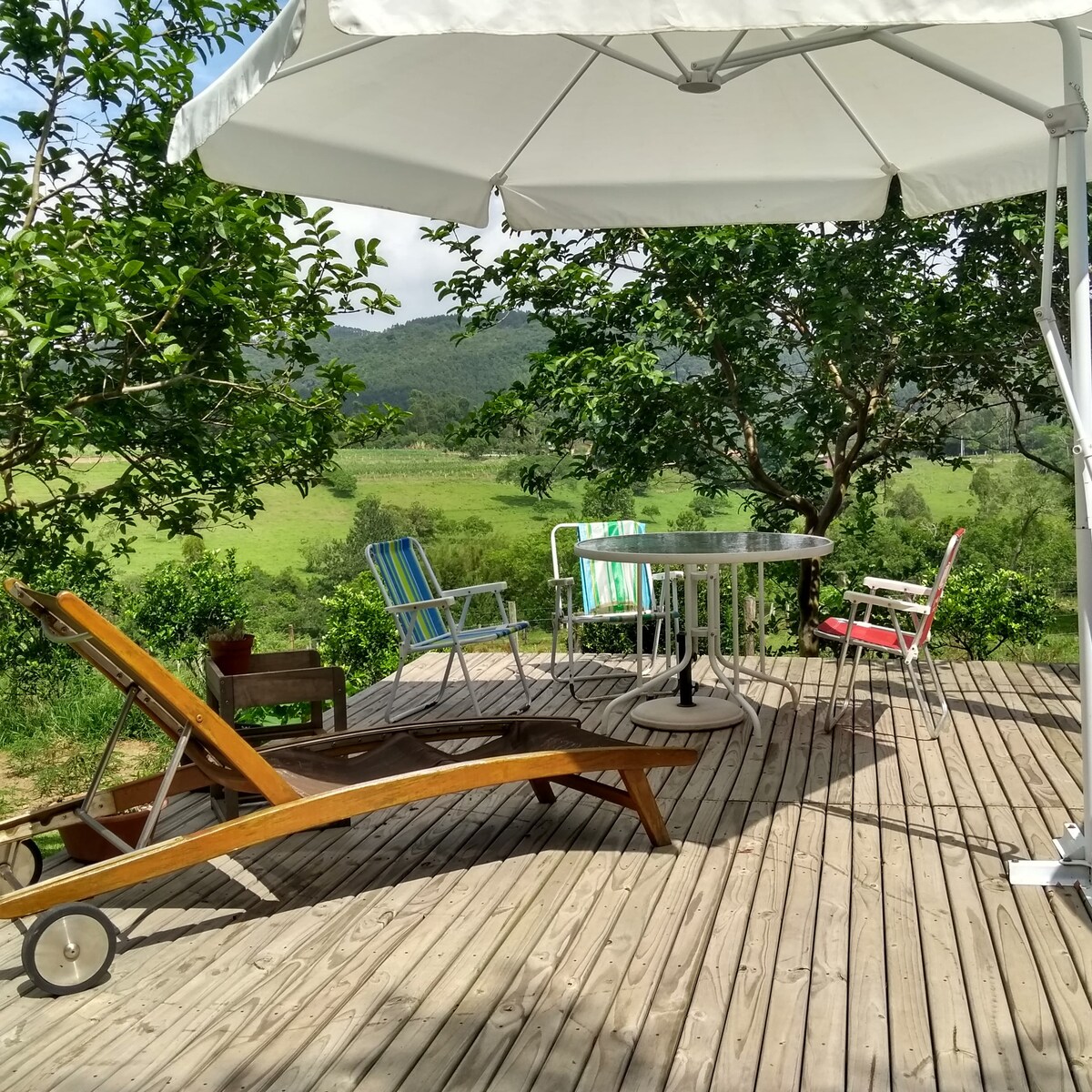 An outdoor deck is furnished with a round table surrounded by colorful chairs. A wooden lounge chair is positioned nearby. The area is shaded by a large umbrella, offering views of green hills and surrounding trees.