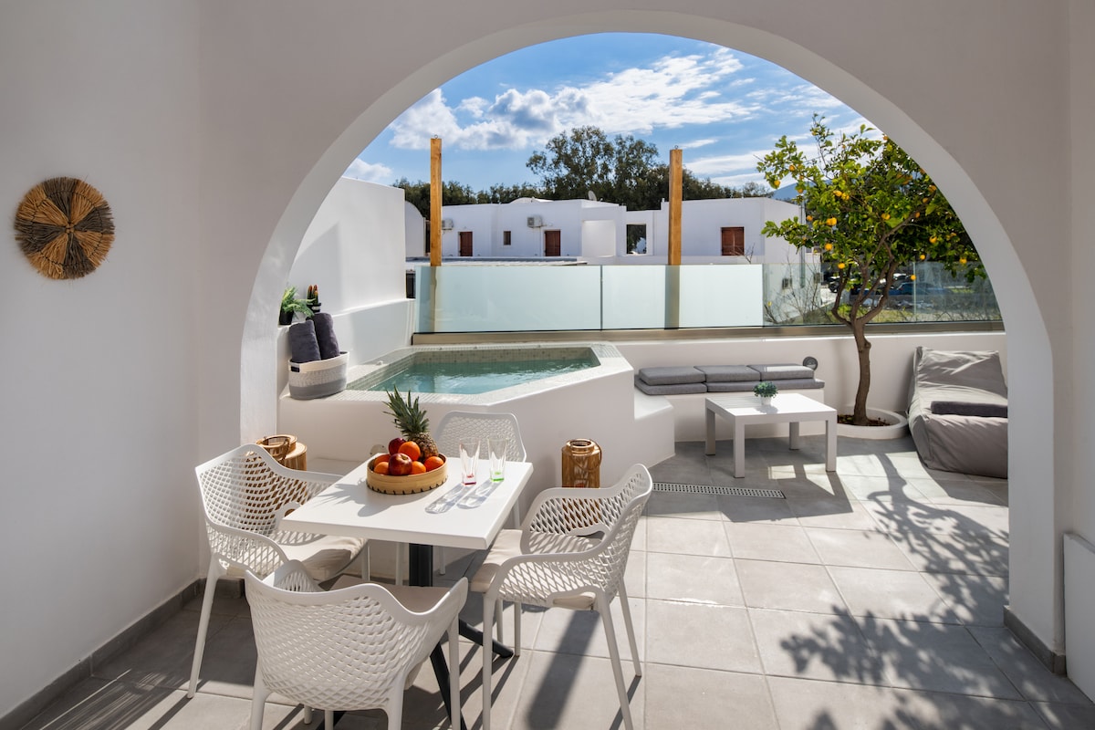 An outdoor space features a heated jacuzzi adjacent to a dining table set for four, surrounded by modern, white chairs. A cozy lounge area with a bench and bean bags is visible, under a bright sky dotted with clouds. Natural light creates a welcoming ambiance.