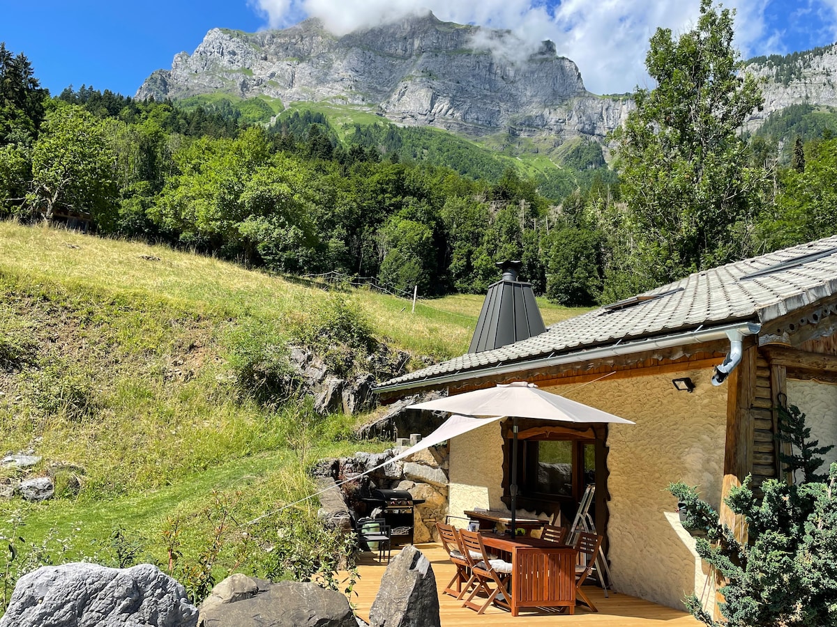 A chalet is positioned near a grassy area, with a mountain in the background. A spacious wooden deck features a table and chairs, shaded by an umbrella. A barbecue grill is visible, creating an inviting outdoor space surrounded by trees.