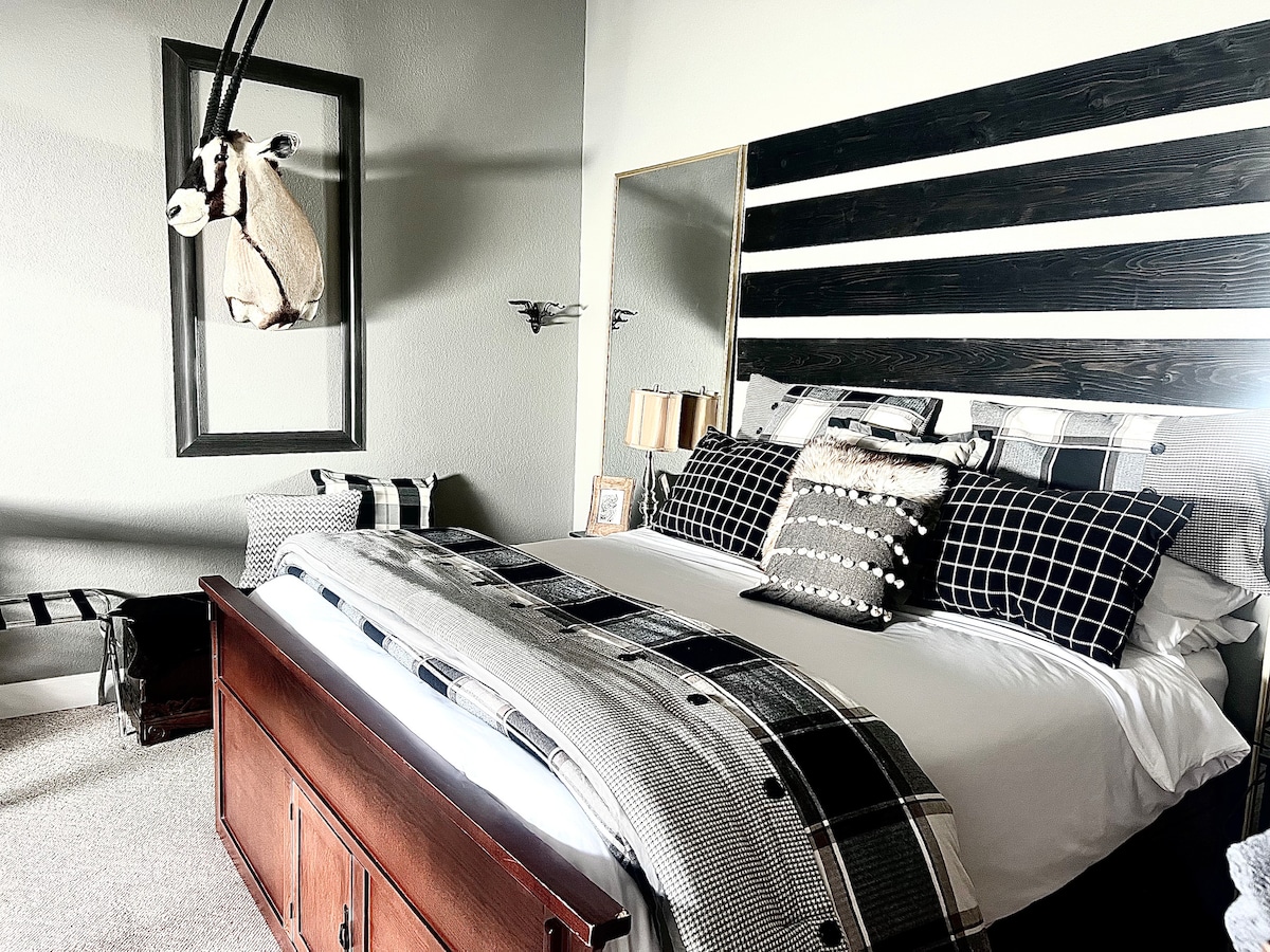 A king bed is positioned prominently, featuring a mix of plaid and solid black and white bedding. Behind the bed, a rustic headboard of horizontal wooden slats is complemented by wall decor. A stuffed animal mount adds a unique touch to the room's design.