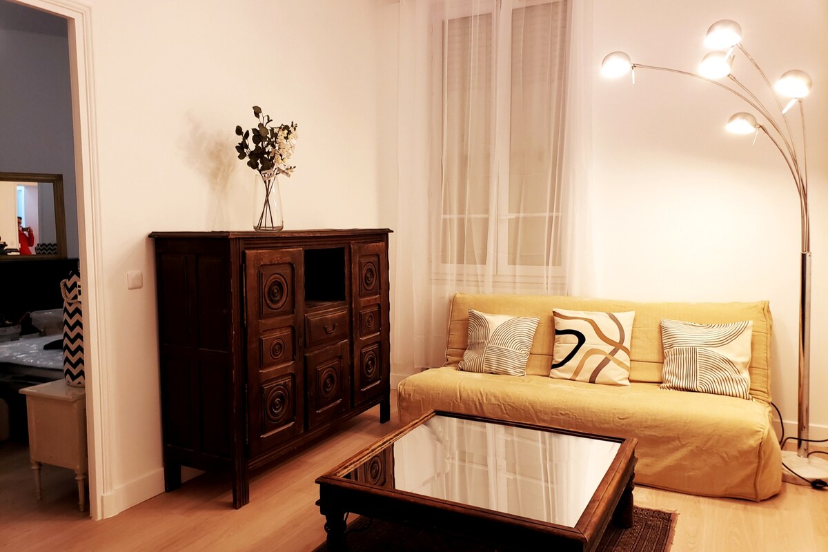 A living room is presented with a yellow sofa adorned with decorative pillows. A mirrored coffee table rests in front of the sofa, while a vintage wooden cabinet adds character to the space. Soft lighting is provided by a modern floor lamp, enhancing the room's ambiance.