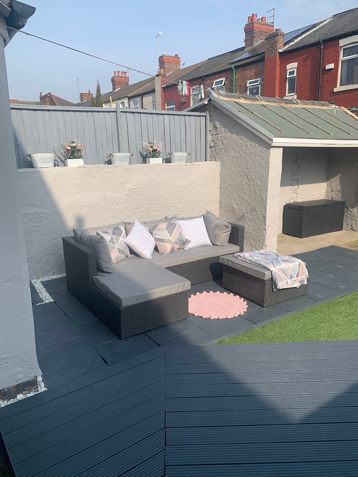 A cozy outdoor seating area is depicted, featuring a sectional sofa adorned with patterned cushions. A soft throw blanket rests over one arm, while a small round rug adds a touch of color. Potted plants enhance the space, providing a refreshing atmosphere.
