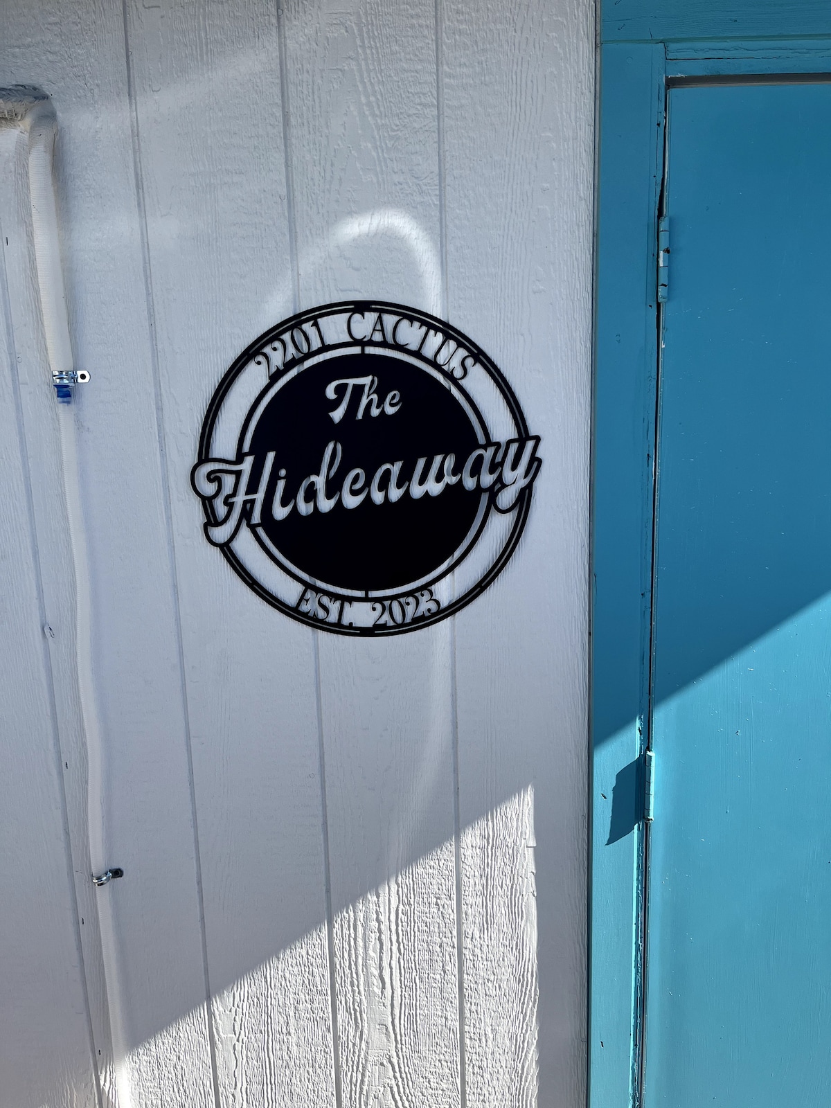 A round sign featuring the name 'The Hideaway' is mounted on a white wall. The sign includes the establishment year 'EST 2023' and is surrounded by a circular black border. A bright blue door can be seen to the side, complementing the sign's colors.