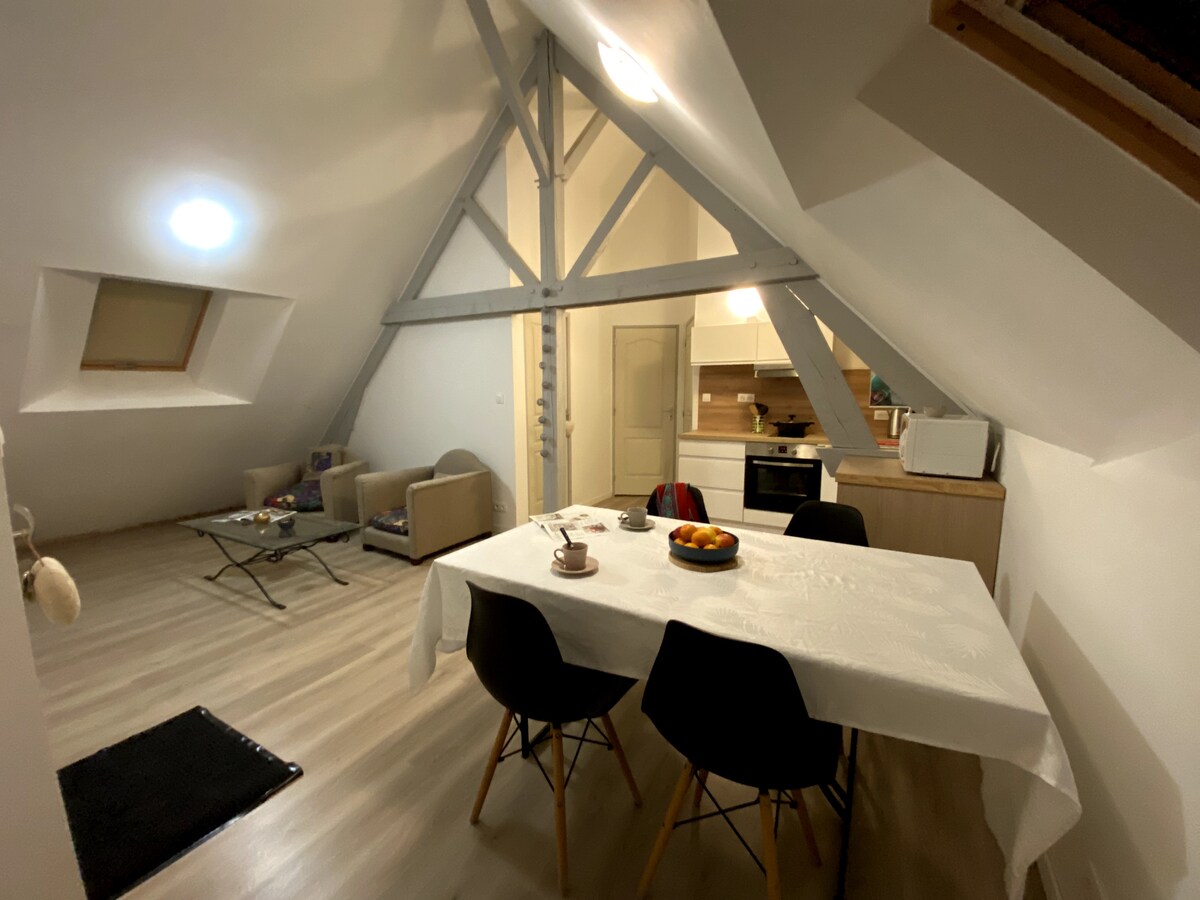 An open living space features a well-lit dining area with a white table set for meals. A cozy seating arrangement is visible in the corner, along with a kitchenette in the background. Modern furnishings and a sloped ceiling add unique character to the room.