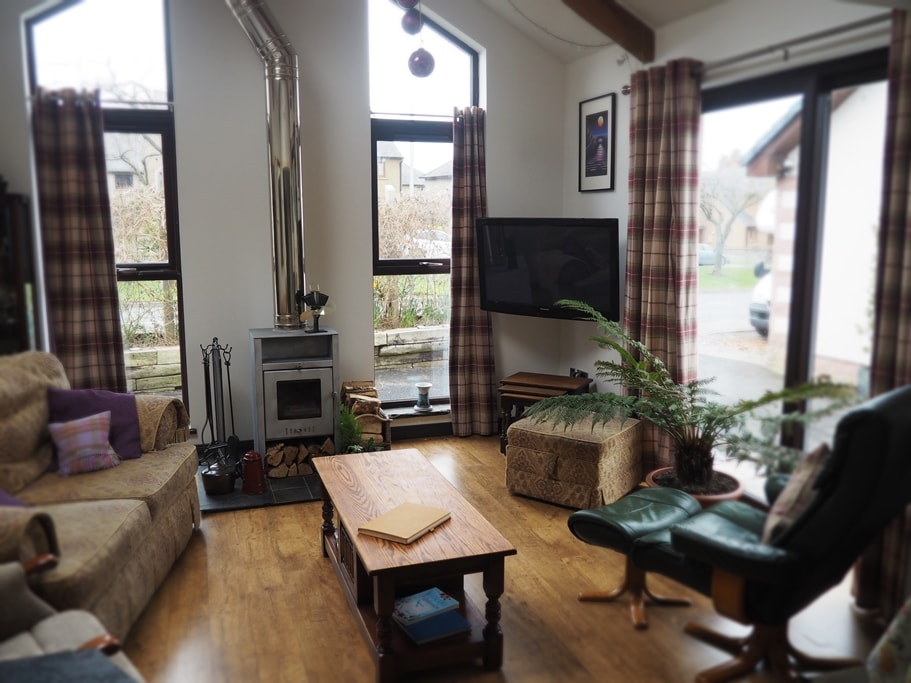 A spacious living area features comfortable seating arrangements, including a green armchair and a beige couch. A wooden coffee table sits in the center, complemented by a flat-screen television mounted on the wall. Natural light enters through large windows, enhancing the room's inviting ambiance.