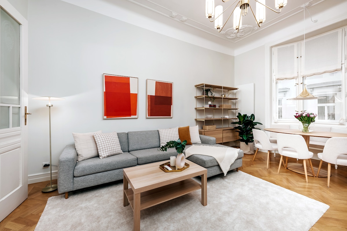 A spacious living area features a light grey sofa adorned with textured cushions, complemented by a wooden coffee table and a soft area rug. Two modern art pieces hang on the walls, and a dining nook with white chairs is positioned near the window, surrounded by greenery.