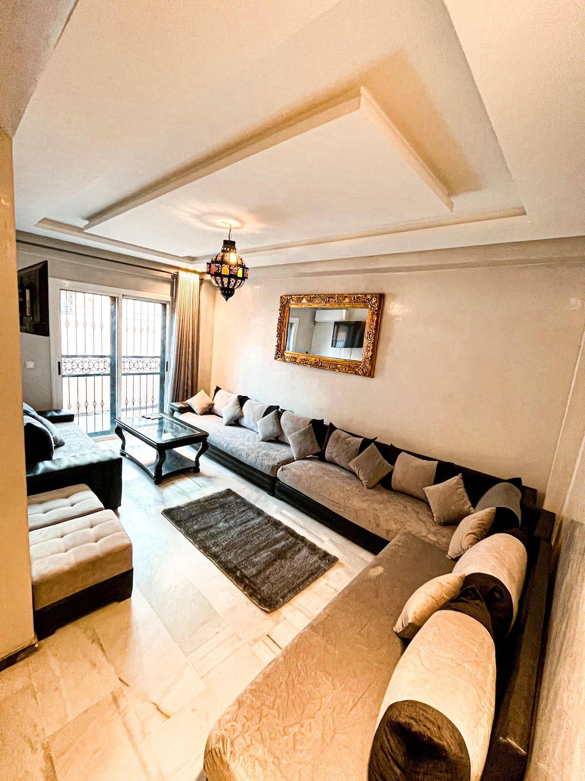 A spacious living area is presented, featuring a large mirrored wall and comfortable L-shaped seating with neutral tones. A central coffee table is surrounded by decorative pillows. Light pours in through large windows, enhancing the serene ambiance of the room.