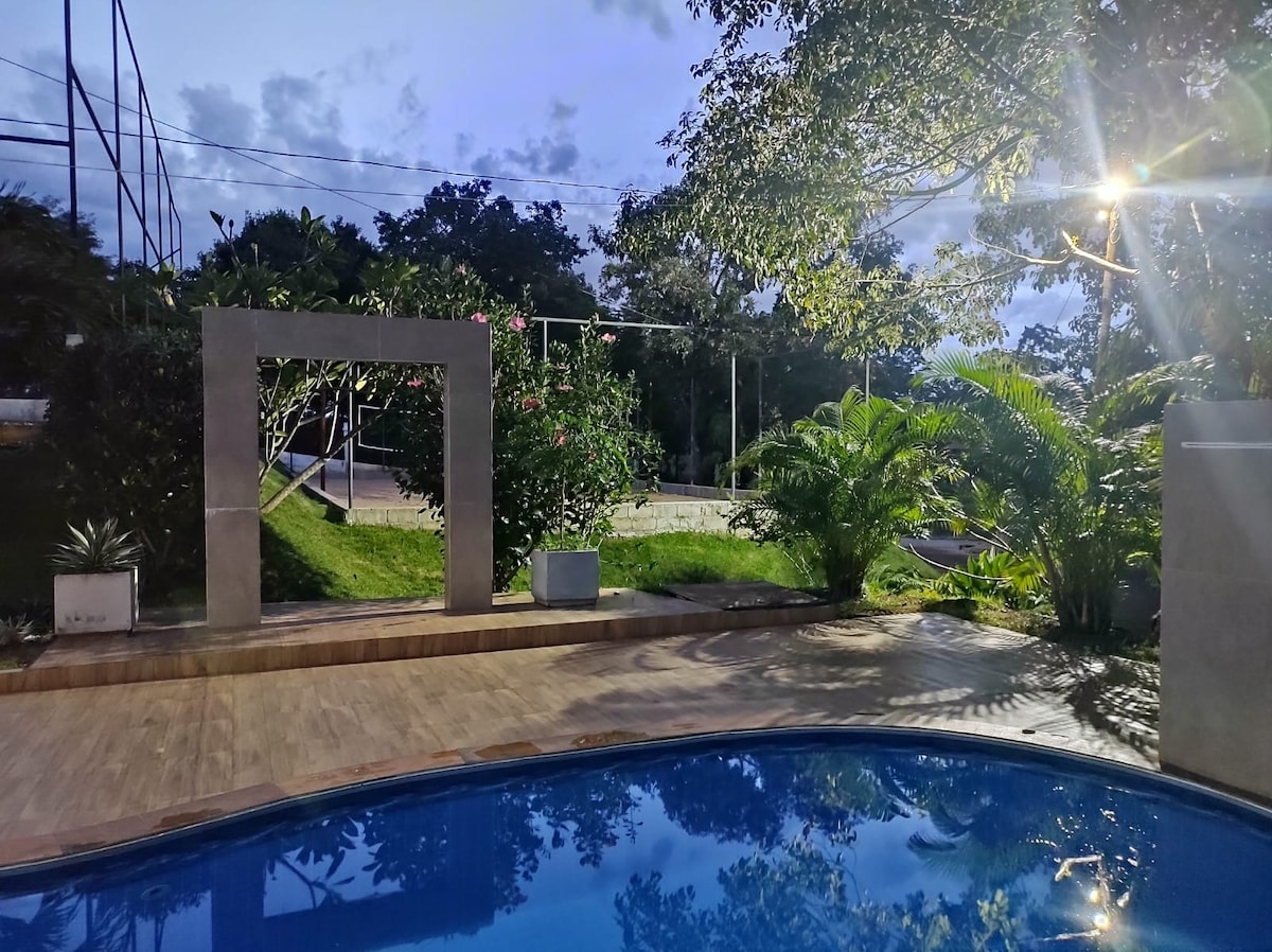 A serene outdoor space is highlighted by a sparkling blue pool, surrounded by lush greenery. In the background, a well-maintained playground area is visible, framed by trees and soft evening lighting, creating an inviting ambiance.