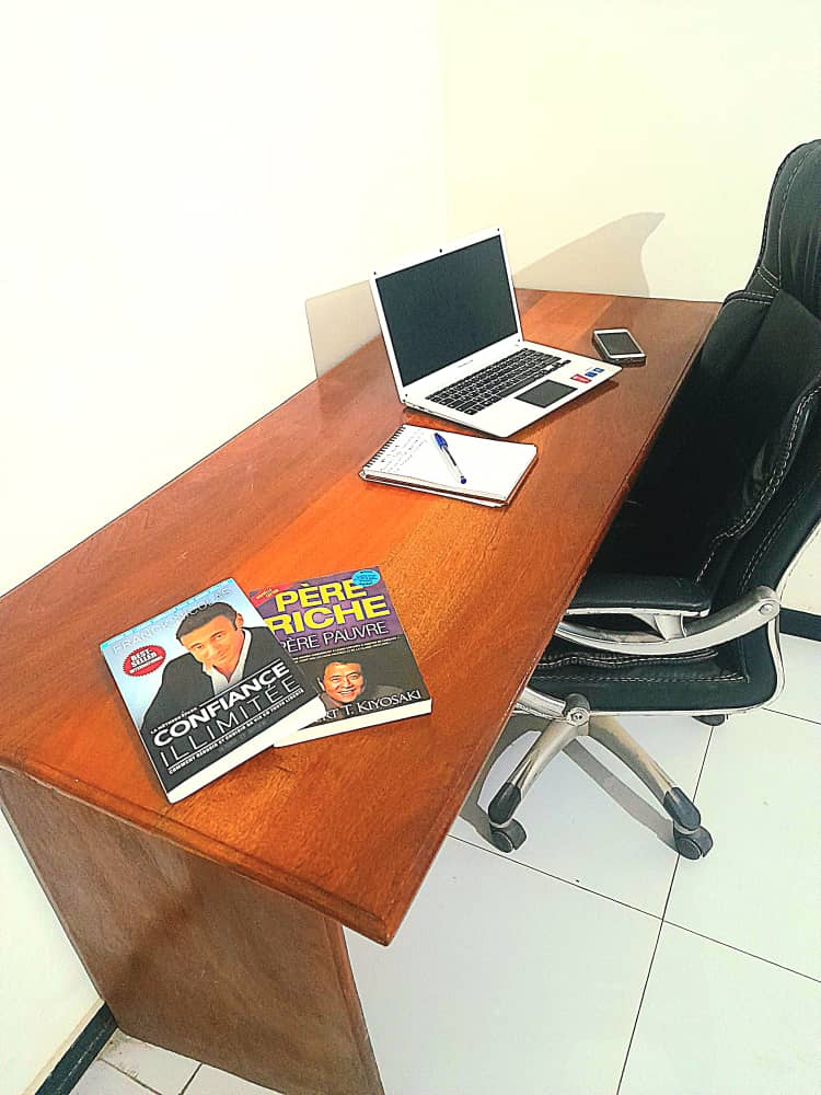 A minimalist workspace is presented, featuring a sleek wooden desk with a laptop and a notepad. A smartphone sits beside the laptop, and two magazines are neatly arranged on the surface. An ergonomic office chair complements the setup, enhancing the productivity-focused environment.