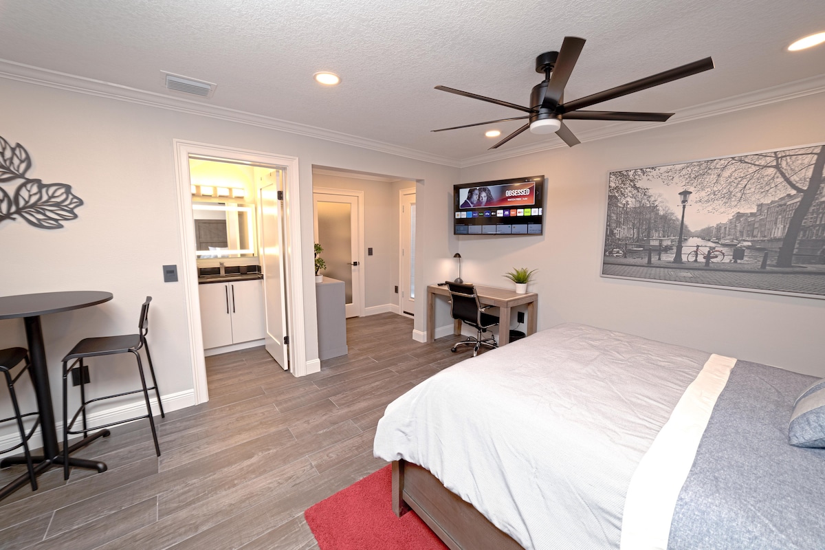 A spacious studio features a queen bed with a light throw and a large window allowing natural light. A ceiling fan is present overhead. A small dining table with two chairs is visible, along with a desk area and a TV mounted on the wall. Access to a bathroom can be seen in the background.