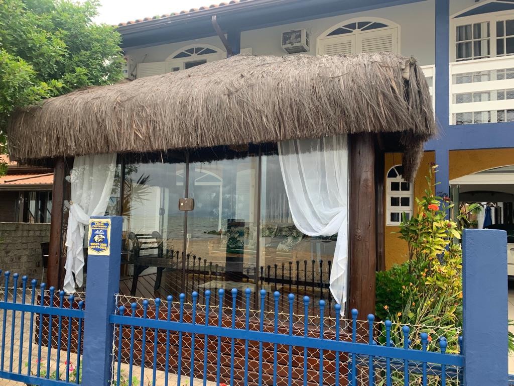 An outdoor seating area is sheltered by a thatched roof, featuring large glass windows that offer views of the surroundings. Lush greenery is present around the space, and light curtains gently frame the windows, creating an inviting and relaxed atmosphere.