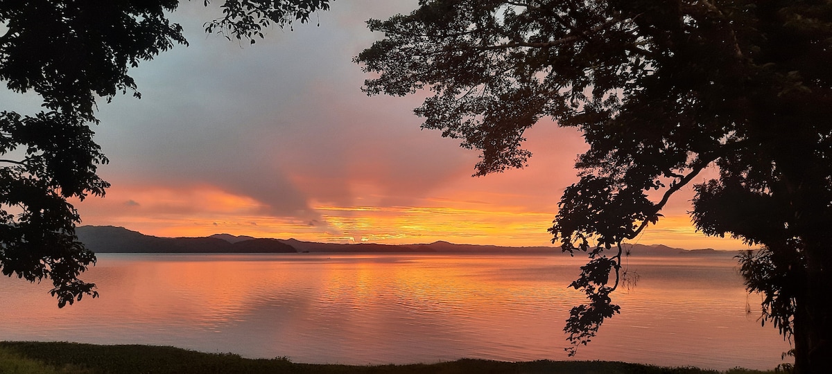 A serene view of a tranquil lagoon at sunset is framed by lush trees. The sky displays a gradient of warm colors, reflecting softly on the water's surface. Silhouettes of distant hills are visible against the vibrant horizon.