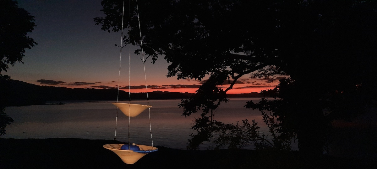 Two hanging bowls are positioned in the foreground against a twilight backdrop. The horizon features soft hues of orange and purple, reflecting over the calm waters of the lagoon. Silhouetted tree branches frame the scene, enhancing the serene atmosphere.