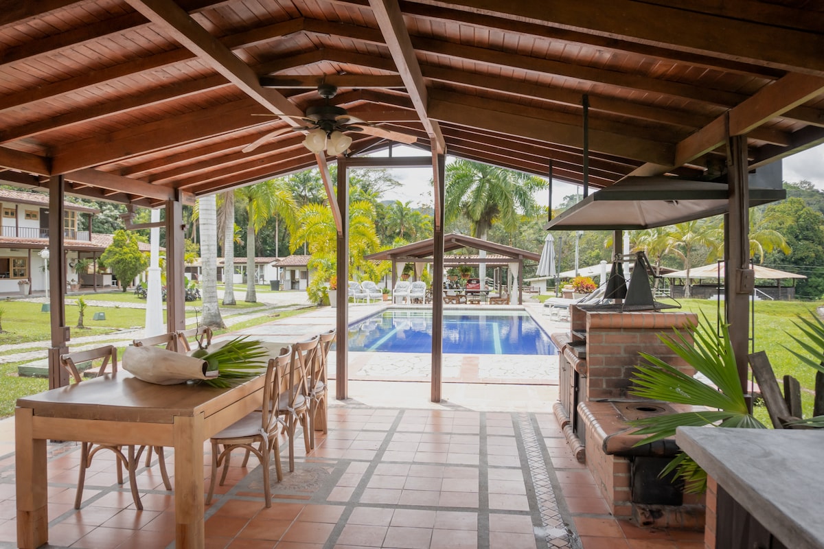 An open dining area features a wooden table surrounded by several chairs. The space is shaded by a wooden roof, offering views of the pool and lush greenery beyond. A barbecue area with a grill complements the inviting outdoor atmosphere.