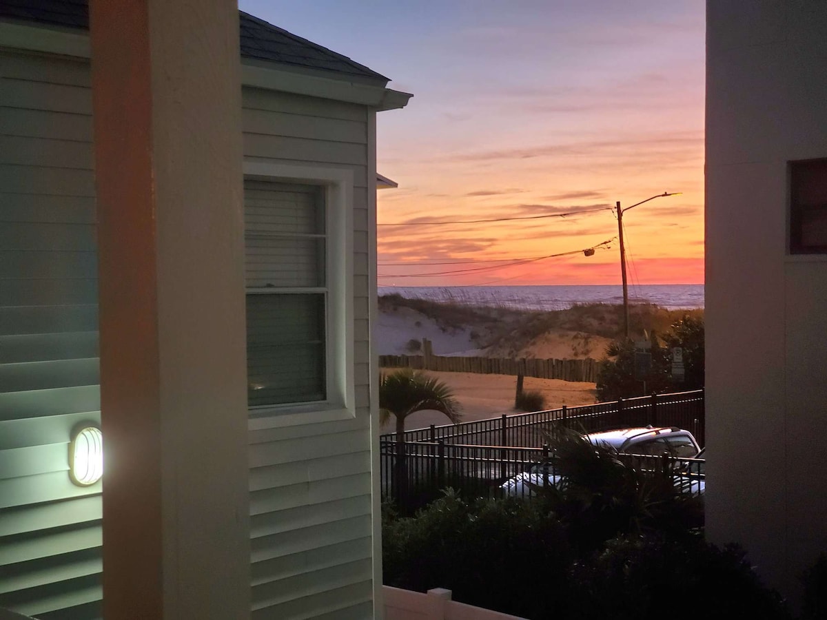 A scenic view is captured at sunset, showcasing vibrant hues in the sky. The beach can be seen in the distance, framed by two buildings. Soft lighting from a wall sconce illuminates the corner of the image.