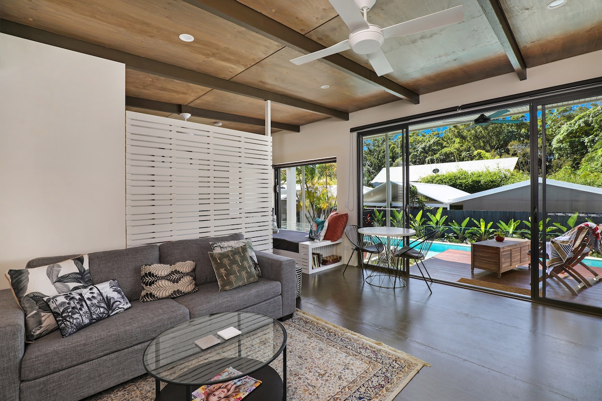 A modern open-plan living area features a grey sofa adorned with decorative pillows, alongside a glass coffee table. Large sliding doors lead to a lush outdoor space with pool views, while a dining area is positioned near the window, complemented by natural light.